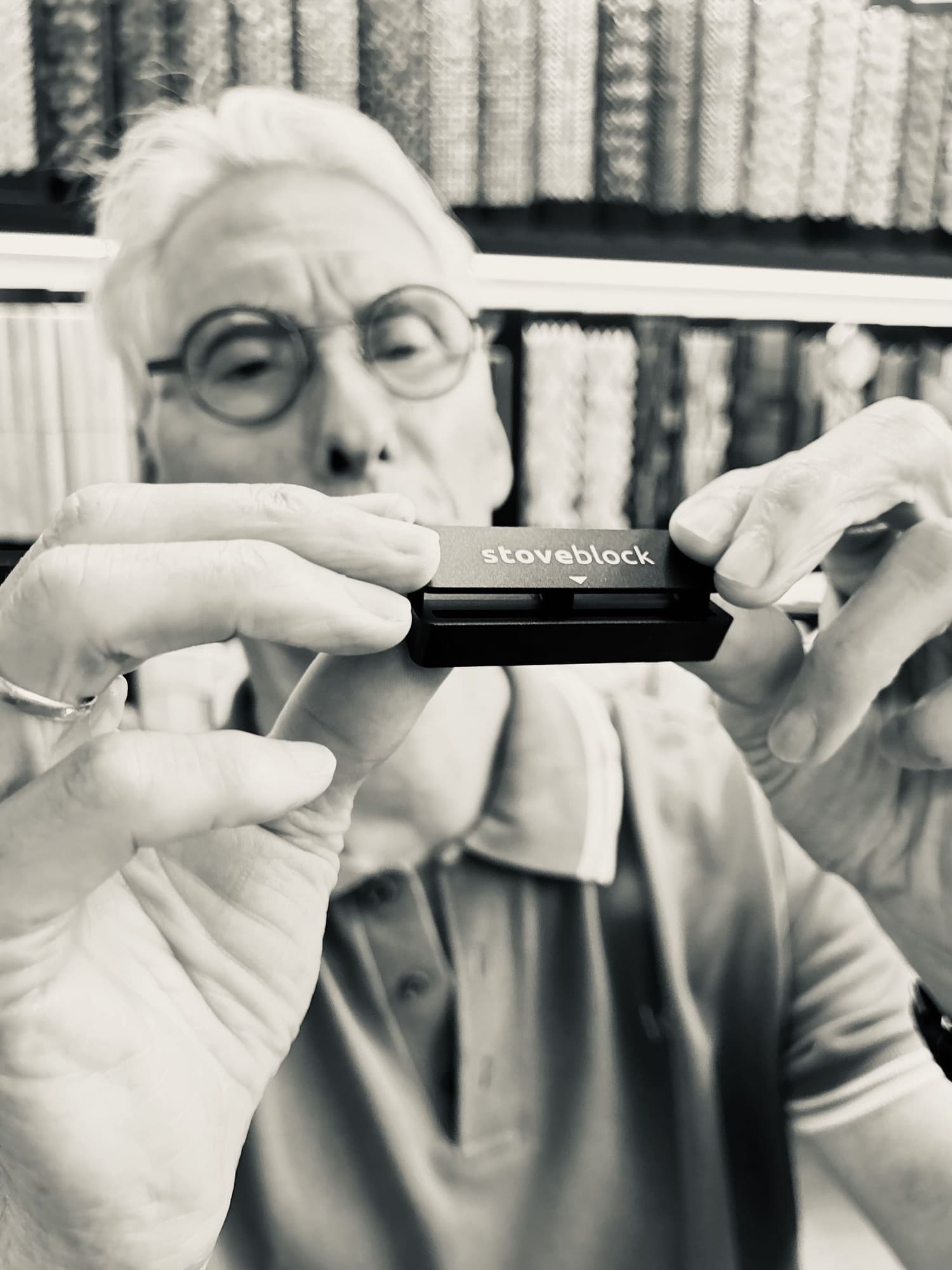 An older man with glasses holds a black Stoveblock stove insert in front of his face, with a blurred background of bookshelves.